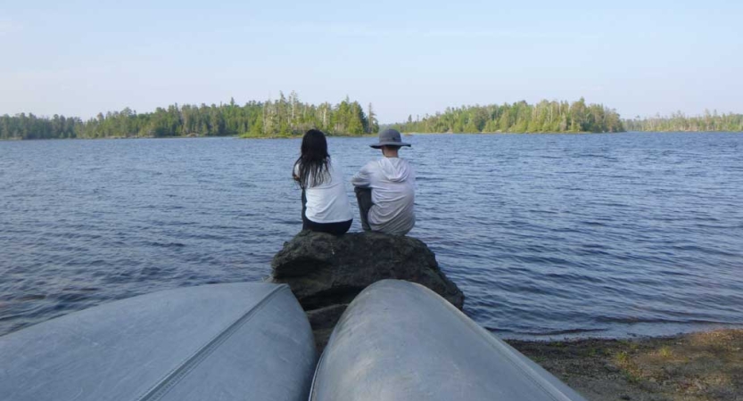 Two people sit on a rock beside a lake shore. Two upside down canoes rest in the foreground.
